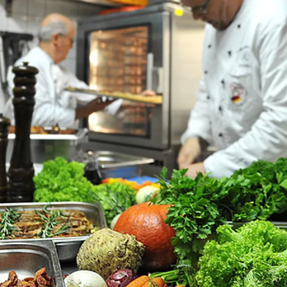 Zwei Köche bereiten in einer Küche frisches Gemüse zu, im Vordergrund Karotten und Salat.