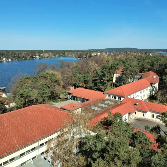 Gebäude mit roten Dächern am See, umgeben von Bäumen unter blauem Himmel.