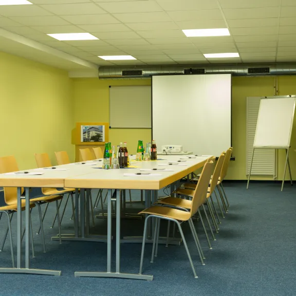 Konferenzraum mit langem Tisch, Stühlen, Leinwand und Flipchart.