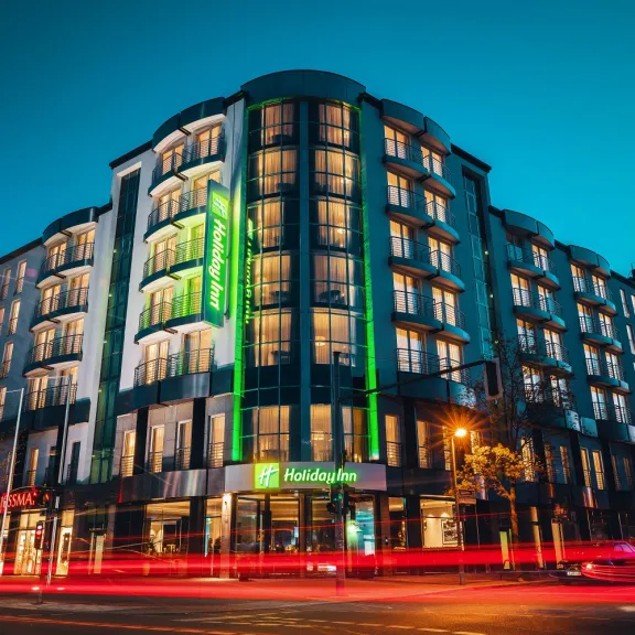 Beleuchtetes Hotelgebäude bei Nacht, vorbeifahrende Autos mit Lichtspuren.