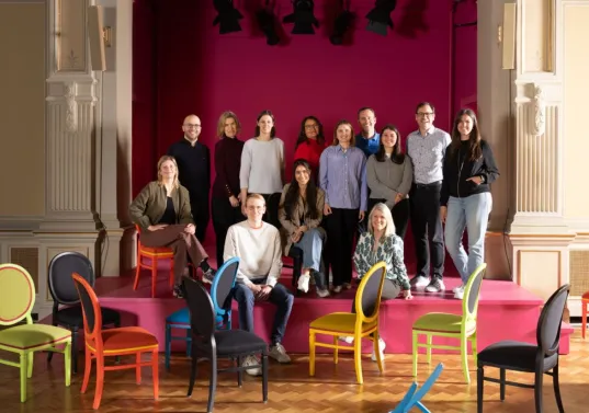Gruppe von Menschen auf einer Bühne, bunte Stühle im Vordergrund, rosa Hintergrund.