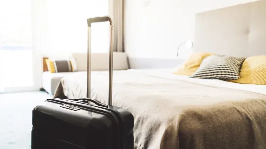 Koffer mit ausgezogenem Griff steht neben einem gemachten Bett in hellem Hotelzimmer.