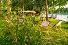 Mehrere Liegestühle stehen auf einer grünen Wiese am Seeufer im Sonnenschein.