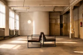 Zwei moderne Sessel stehen Rücken an Rücken in leerer, lichtdurchfluteter Industriehalle.