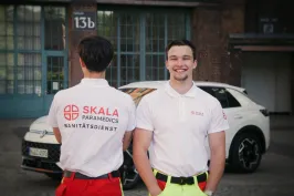 Zwei Rettungskräfte von Skala Paramedics in weißer Dienstkleidung vor einem weißen Einsatzfahrzeug; eine Person ist von hinten zu sehen, die andere lächelt in die Kamera.