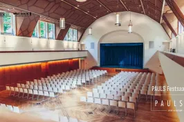 Großer, heller Saal mit Reihen weißer Stühle und Bühne mit blauem Vorhang.