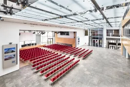 Großer, heller Hörsaal mit vielen roten Stuhlreihen und großer Fensterfront.