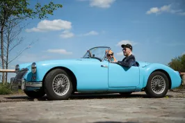 Mann mit Hund fährt bei Sonnenschein in einem blauen Cabrio über Kopfsteinpflaster.