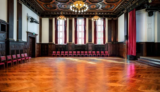 Großer Saal mit Holzfußboden, hohen Fenstern und Stuhlreihe an der Wand
