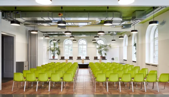 Konferenzraum mit grünen Stühlen, Bühne vorne, große Fenster, helle und moderne Atmosphäre.
