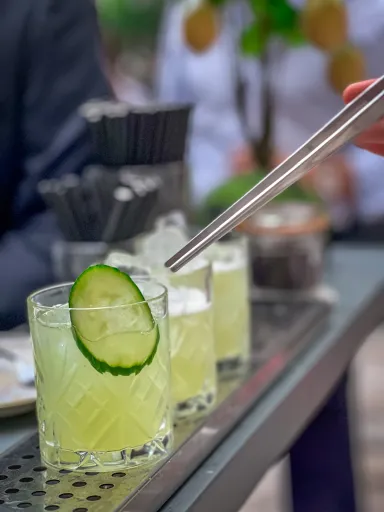 Glas mit Gurkenscheibe und Cocktail auf einer Bar, Person hält einen Metallstrohhalm.