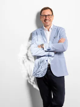 Mann mit Brille und hellem Sakko steht lächelnd vor weißer Wand.