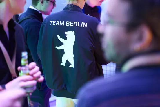 Person mit Jacke "TEAM BERLIN" und Bärensymbol, umgeben von mehreren Menschen.