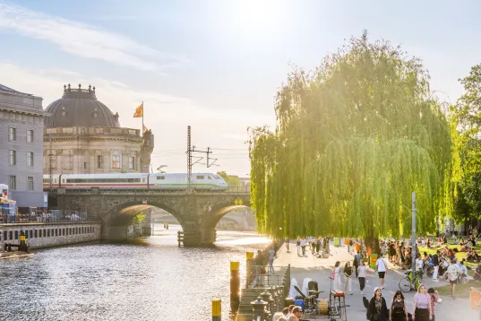 Sonniger Tag an der Spree mit Brücke, Zug und vielen Menschen am Ufer.