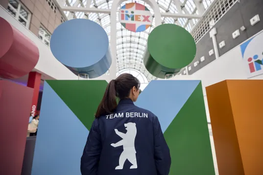 Frau mit „Team Berlin“-Jacke vor bunten Skulpturen in einer Messehalle.
